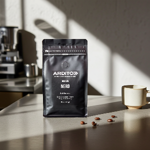 A black stand-up pouch of ARDITO Fuel coffee labeled 'Riserva Nero' stands on a countertop in a kitchen setting, with coffee beans scattered nearby and a ceramic cup to the right. The scene is lit by strong natural sunlight with soft shadows.