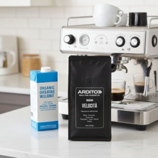 A black bag of ARDITO Velocita espresso beans stands on a white countertop next to a blue and white carton of Organic Orgaine Velorif milk, with a stainless steel espresso machine in a bright kitchen background.